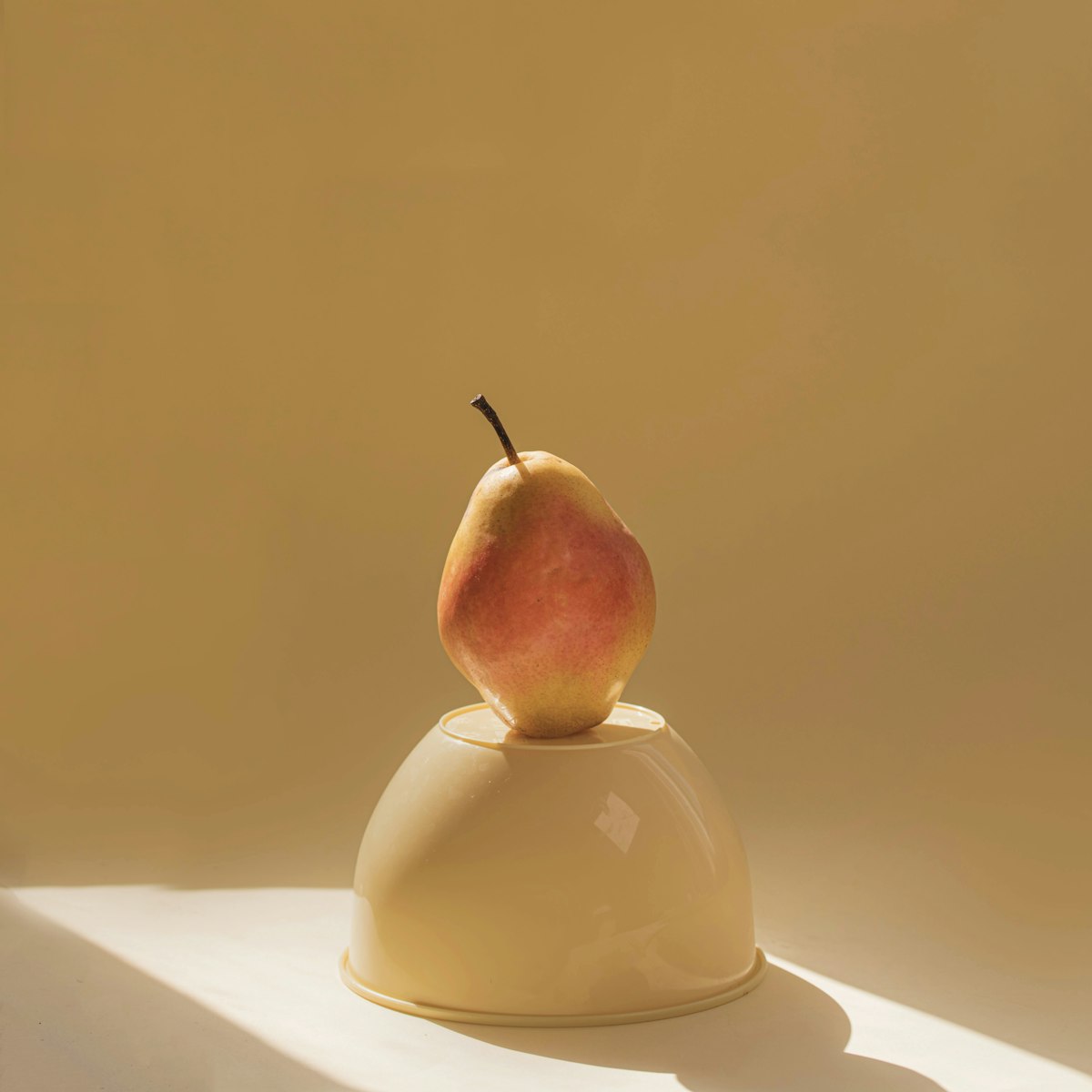 A cream bowl turned over with a pear on top, against a mustard wall
